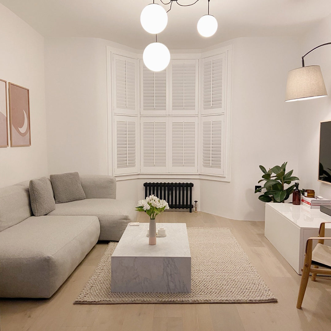 A modern living room featuring a light gray L-shaped sectional sofa, a white marble coffee table, a textured beige rug, and a white bay window with shutters in the background.