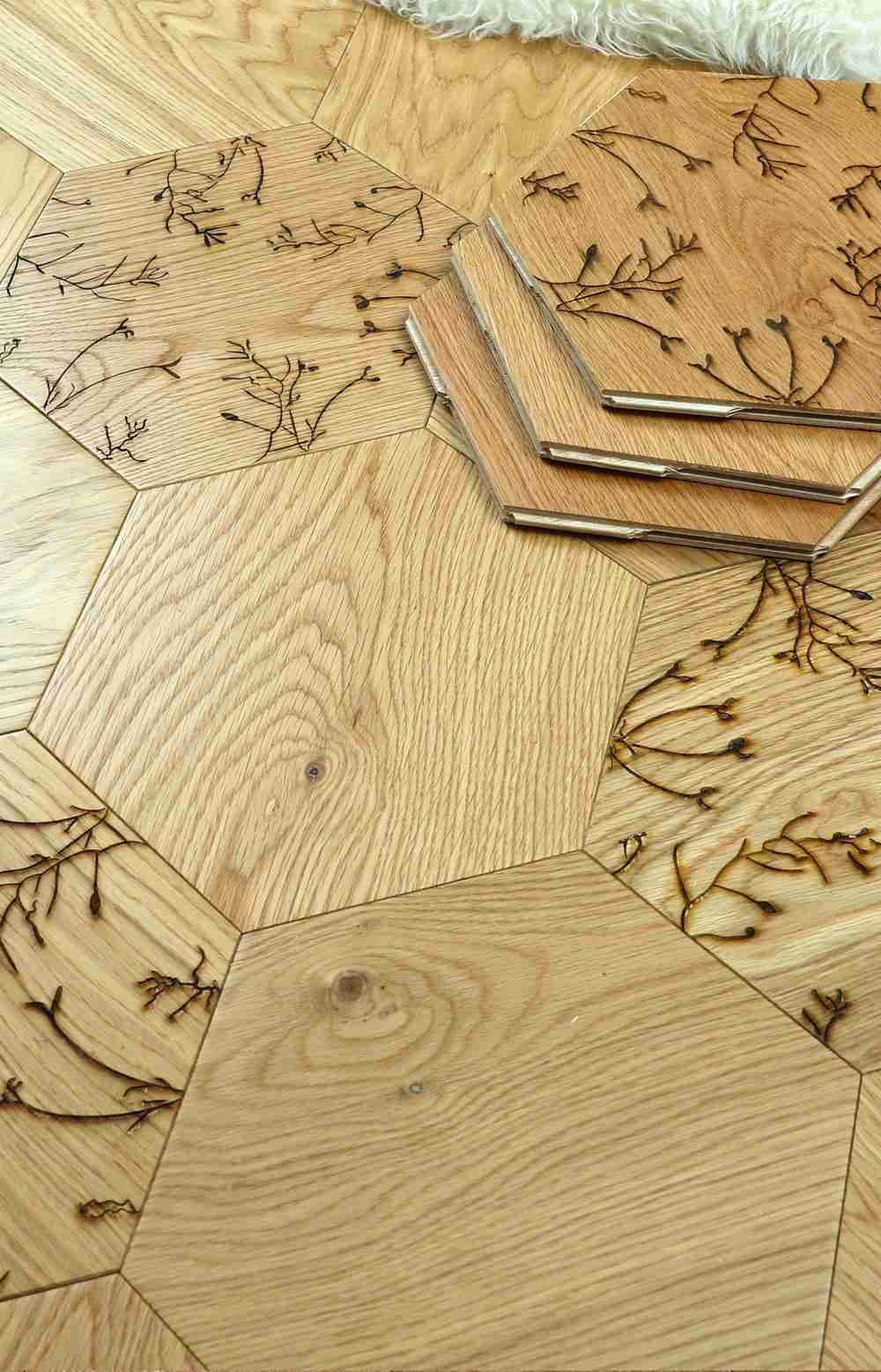 Hexagonal wood flooring tiles with natural grain and laser-burned botanical patterns, shown with a stack of sample pieces on a white rug.