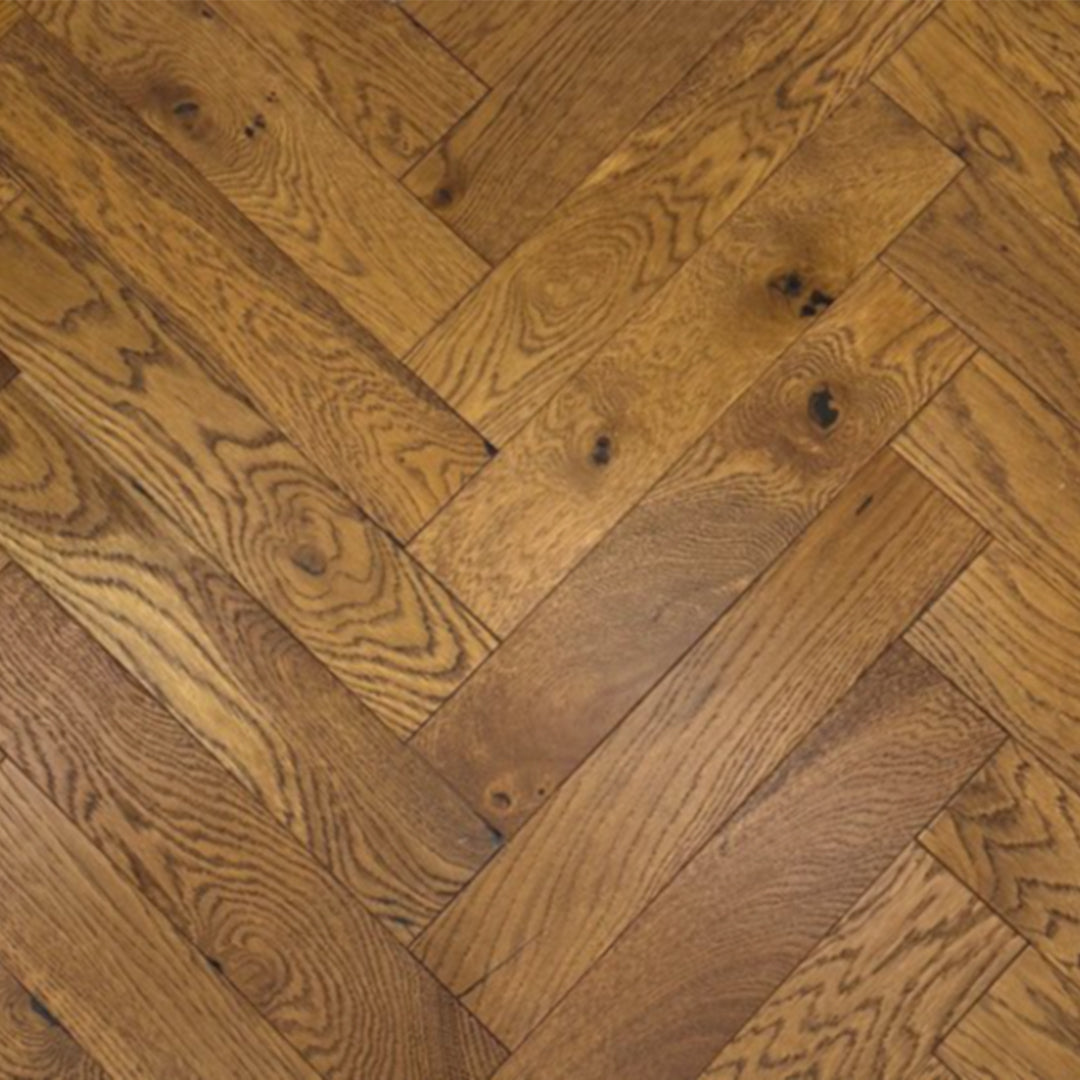 A close-up view of a herringbone parquet wood floor with warm medium-brown tones and visible natural grain patterns and knots.