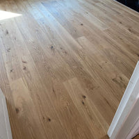 Light oak hardwood flooring planks with visible natural grain, knots, and warm tan tones, shown installed in an interior space with white trim visible on the right and bottom edges.