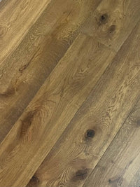 Natural oak hardwood flooring planks with visible grain, knots, and warm medium-brown tones arranged diagonally across the frame.