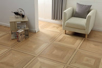 Light wood geometric parquet flooring installed in a modern living room setting, shown with a cream armchair, a small wooden side table, and a gray curtain.
